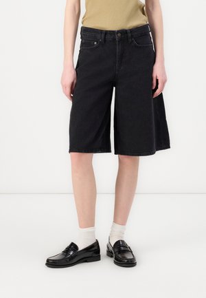 Shorts en denim noir avec une coupe décontractée, longueur genou, design à cinq poches et détails de couture visibles. Associés à des mocassins noirs et des chaussettes blanches.