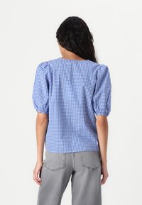 Blå og hvit gingham-rutete bluse med puffede ermer og rund hals. Stoffet virker jevnt med en lett tekstur. Brukt med grå jeans.