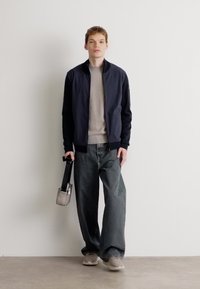 Námornícky bomber jacket, šedý pletený sveter, široké šedé džínsy, svetlosivé tenisky, drží malú šedú tašku. Hladká tkanina a voľný strih.