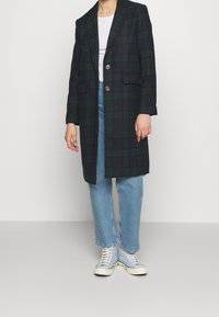 Kabát z kostkované vlny v tmavě zelené a námořnické modři, s klopovým límcem, dvěma předními kapsami a dvěma knoflíky, nošený přes světle modré džíny a tenisky.