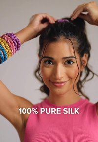 Vaaleanpunainen ribbitekstiilitoppia, joka on valmistettu 100 % puhtaasta silkkistä. Kirkkaan värisiä teksturoituja hiuslenkkejä, jotka ovat väriltään violetteja, vaaleanpunaisia ja sinisiä, käytetään ranteessa. Neutraali tausta.