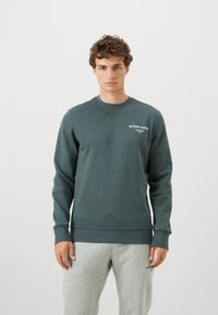 Nuori mies, jolla on kihara hius, pukeutunut tumman teal Björn Borg -sweatshirthiin ja vaaleanharmaisiin collegehousuihin yksinkertaista vaaleaa taustaa vasten.