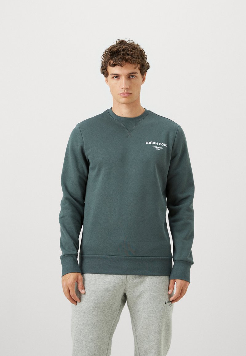 Nuori mies, jolla on kihara hius, pukeutunut tumman teal Björn Borg -sweatshirthiin ja vaaleanharmaisiin collegehousuihin yksinkertaista vaaleaa taustaa vasten.