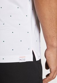 Bílý sportovní tričko s zeleným čtvercovým vzorem, krátkými rukávy, bočními rozparky a štítkem s logem na spodním lemu. Materiál vypadá hladce a prodyšně.