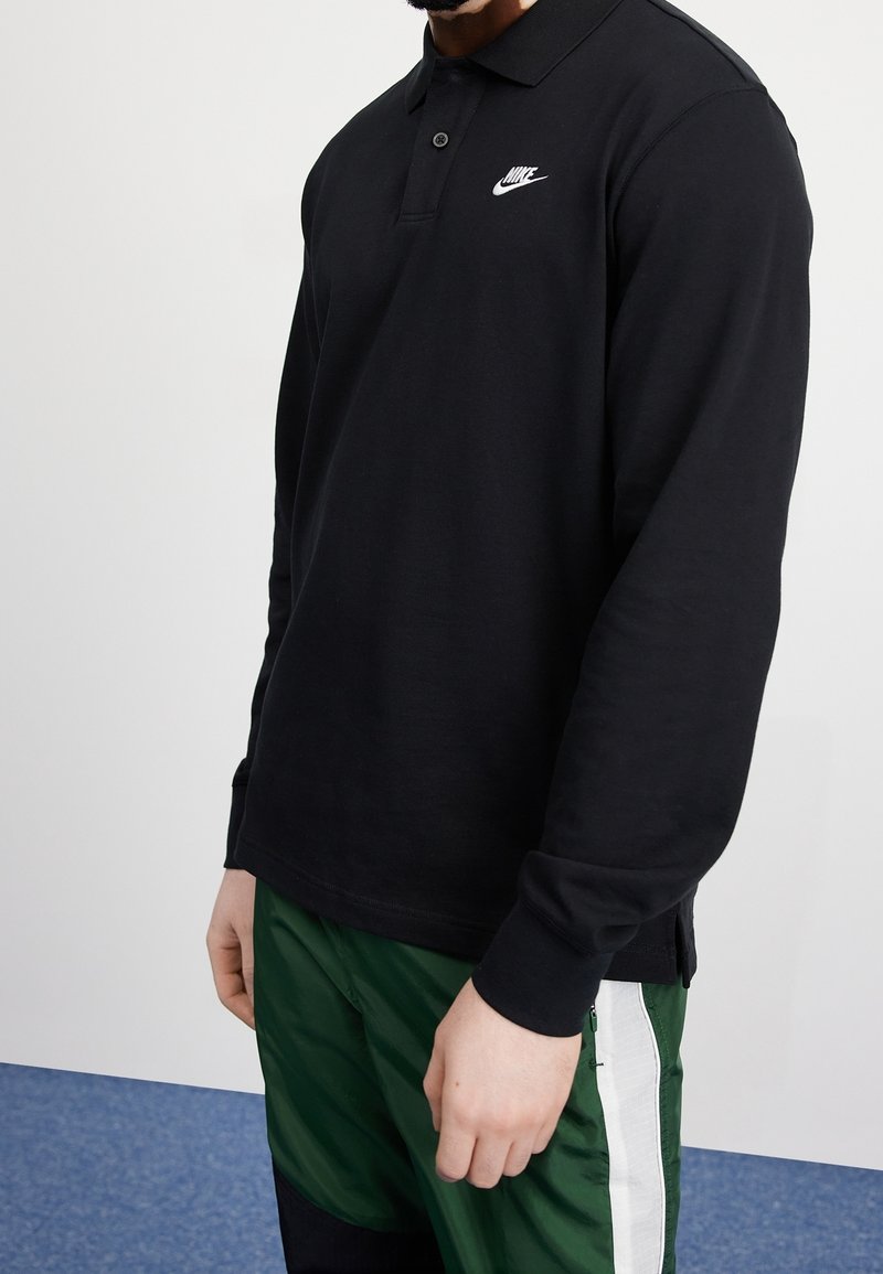 Mężczyzna w czarnej koszulce polo Nike z długim rękawem i zielonych spodniach dresowych z białym paskiem, stojący na tle białym i niebieskim.