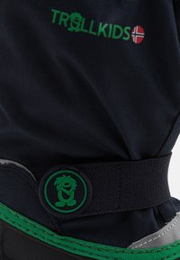 Jūras spēku auduma zābaki ar zaļu elastīgu siksnu un apļa logotipu, sudraba akcentiem un teksturētu virsmu. Zīmols ir redzams augšpusē.