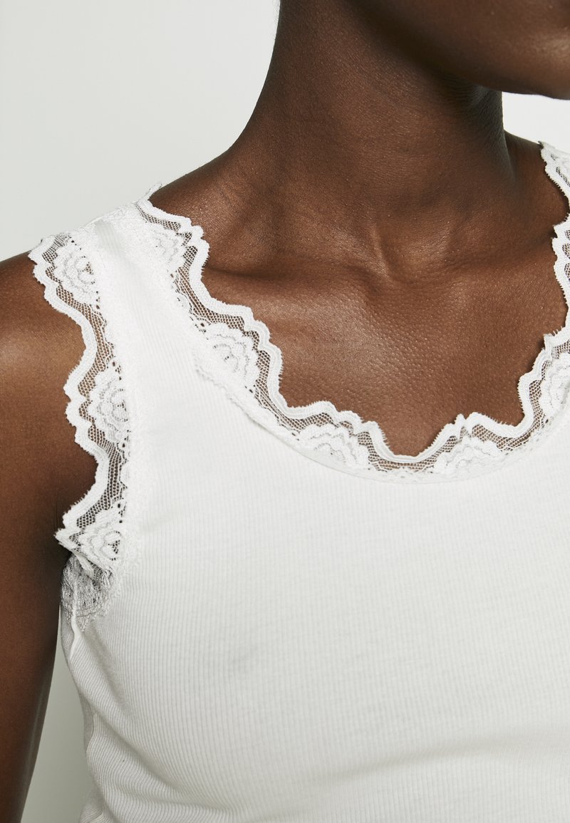 Gros plan d'une personne à la peau foncée portant un haut blanc sans manches avec un bord en dentelle festonné autour de l'encolure et des emmanchures.
