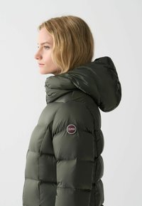 Olivgrön pufferjacka med avtagbar huva, med en rund logotyp på vänster axel och kviltad design för isolering.