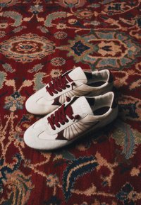Paire de baskets blanches avec des lacets rouge foncé sur un tapis détaillé rouge, bleu et beige à motifs.