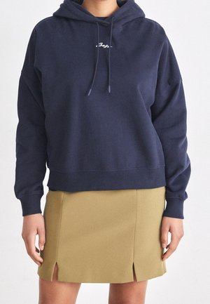 Personne portant un sweat à capuche bleu marine avec du texte blanc et une minijupe beige avec deux petites fentes à l'avant.