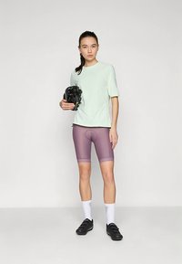 Jasnozielony top z krótkim rękawem w połączeniu z fioletowymi szortami kolarskimi; szorty mają obcisły krój i subtelny wzór, noszone z czarnymi butami kolarskimi.