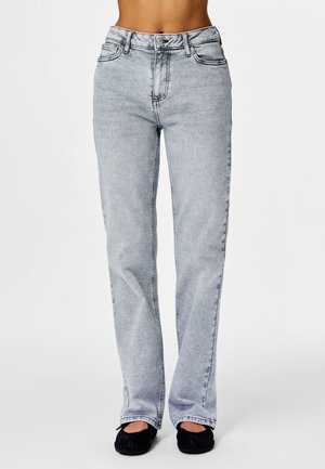 Persona, kas valkā gaiši pelēkas taisnas bikses ar augstu jostasvietu un melnus dzīvokļveida apavus, stāvot pret vienkāršu baltu fonu.