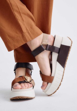 Pies con sandalias de plataforma con tiras anchas, texturizadas y marrones multicolores, combinadas con pantalones cortos de color naranja quemado, de pie sobre un suelo blanco.