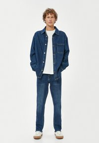 Chaqueta de denim y jeans en azul oscuro. La chaqueta cuenta con un bolsillo frontal y cierre de botones, combinada con una camiseta blanca y zapatos blancos con suelas marrones.