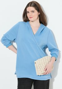 Světle modrá mikina s kapucí a v-neck výstřihem, dlouhými rukávy a oversized střihem. Texturovaná krémová kabelka s bobble vzorem držená v ruce.