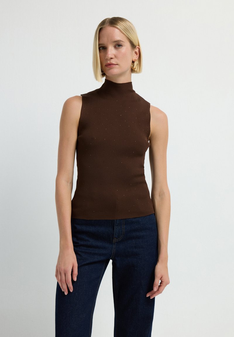 Vrouw met kort blond haar, gekleed in een mouwloze ribgebreide bruine coltrui en donkerblauwe jeans, staand tegen een effen lichte achtergrond.