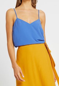 Una donna indossa un top blu senza maniche con spalline sottili e una gonna avvolgente giallo senape a vita alta con una cintura laterale.