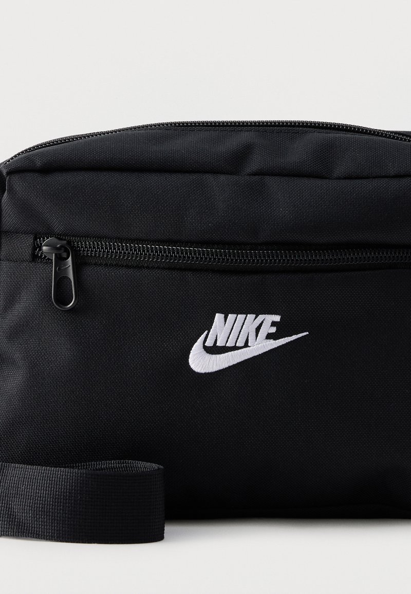 Schwarze Nike Bauchtasche mit vorderer Reißverschlusstasche und verstellbarem Riemen, mit weiß gesticktem Nike-Logo und Swoosh.