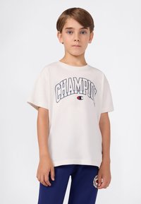 Chłopiec z krótkimi brązowymi włosami, ubrany w białą koszulkę Champion i granatowe spodnie z naszywką logo, stojący na jednolitym tle.