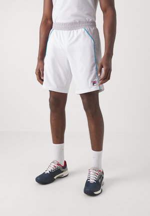 Hombre con pantalones cortos deportivos blancos con cintura gris y ribete azul, calcetines blancos hasta el tobillo y zapatillas FILA azul marino y rojo.