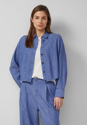 Jeune femme portant une veste-chemise bleue boutonnée sur un haut blanc et un pantalon taille haute assorti bleu, sur un fond gris.