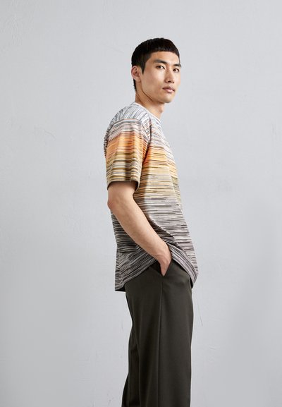 Striped T-shirt featuring horizontal lines in orange, yellow, and gray. Crew neck and short sleeves. Worn with dark, tapered trousers.