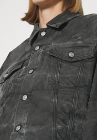 Svart denimjacka med en tvättad yta, med två bröstfickor, stora knappar och en spetsig krage.