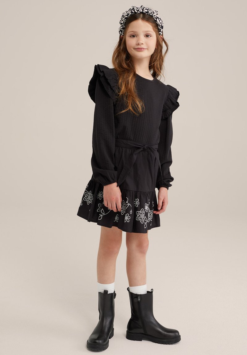 Zwarte jurk met ruches aan de mouwen en bloem borduursel aan de zoom, gecombineerd met kniehoge zwarte laarzen en witte enkelsokken.