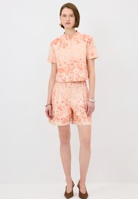Haut à manches courtes de couleur corail avec un motif floral et un col montant, accompagné d'un short assorti, fabriqué en tissu texturé. Chaussures à talons marron.