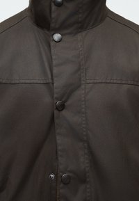 Σκούρο καφέ τζάκετ μπροστινή όψη με ψηλό γιακά, εμφανείς ραφές, κουμπιά με πατιλέτα και φερμουάρ που καλύπτεται εν μέρει από υφασμάτινη πτερύγιο.