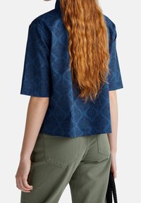 Camicia navy con maniche corte, motivo a rombi, tessuto testurizzato e design cropped, abbinata a pantaloni verde oliva.