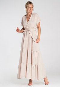 Maxi sukienka z miękkiego beżowego materiału, z kołnierzykiem, zapięciem na guziki, talią ściągniętą w pasie i warstwowym dołem. Posiada pasek w talii oraz krótkie rękawy.