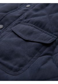 Giacca trapuntata navy con texture morbida, dotata di una tasca a patta e bottoni neri tondi. Cuciture visibili e motivo a diamante sottile.