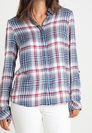 Woman wearing a long-sleeve plaid shirt with blue, red, and white checks, paired with white pants, against a plain background.