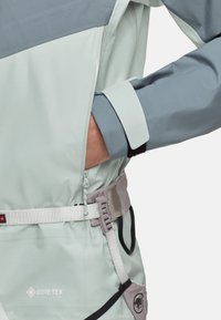 Lys grå jakke med et mørkere grå panel. Har en sidelomme med lynlås, justerbare manchetter og et GORE-TEX-logo. Inkluderer et bælte med hardware.