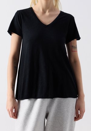Vrouw draagt een losse zwarte V-hals T-shirt met korte mouwen en lichtgrijze broek, toont tatoeages op beide onderarmen tegen een effen achtergrond.