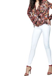 Femme portant un chemisier à motifs de papillon, un jean skinny blanc et des sandales à talons rouges, se tenant les mains sur les hanches.