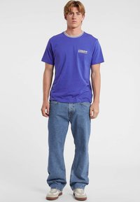 Młody mężczyzna stoi, ubrany w fioletowy t-shirt "Guess Jeans", niebieskie dżinsy i białe trampki, z neutralnym wyrazem twarzy, na jednolitym białym tle.