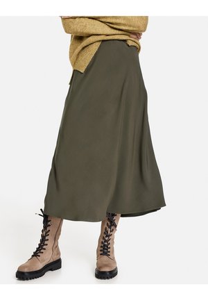 Personne portant une longue jupe vert olive, des bottes de combat beiges à lacets et un pull en maille moutarde, les bras croisés.
