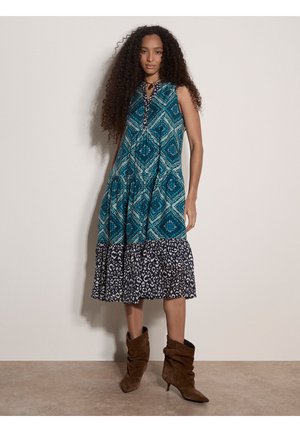 Frau mit langen lockigen Haaren, die ein ärmelloses blau gemustertes Midikleid und braune, lässige Knöchelstiefel trägt, steht vor einem schlichten Hintergrund.