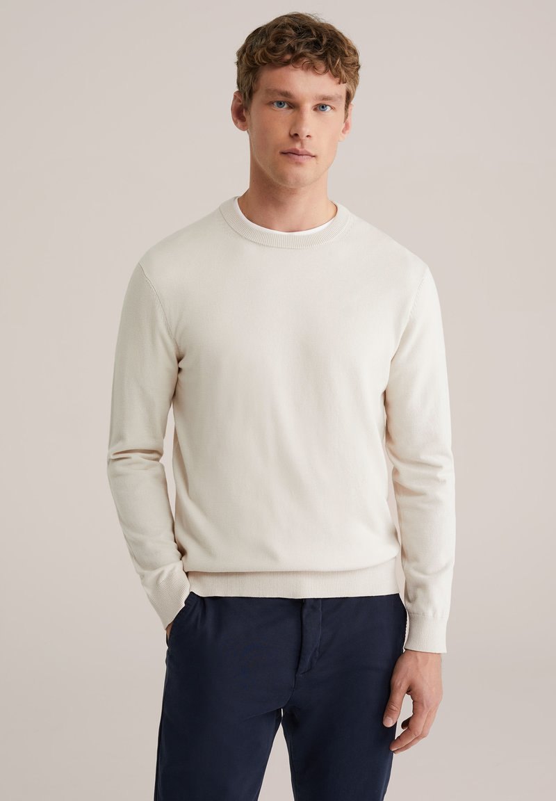 Pull col rond beige clair en tissu tricoté doux, avec poignets et ourlet côtelés, porté avec un pantalon bleu marine foncé.