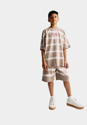 Beige Batik-T-Shirt mit kurzen Ärmeln und passender Shorts, mit einem zentrierten roten Textdesign. Weiße Sneakers mit cremefarbenen Sohlen.