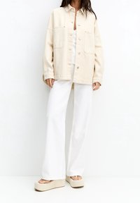 Beige oversized denim jack met twee voorzakken, knoopsluiting en relaxte pasvorm, gecombineerd met witte wijde broek en platform sandalen.