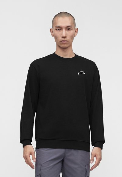 Homme à la tête rasée de près portant un sweat-shirt à manches longues noir avec un petit logo blanc sur la poitrine et un pantalon cargo gris, debout devant un fond blanc.