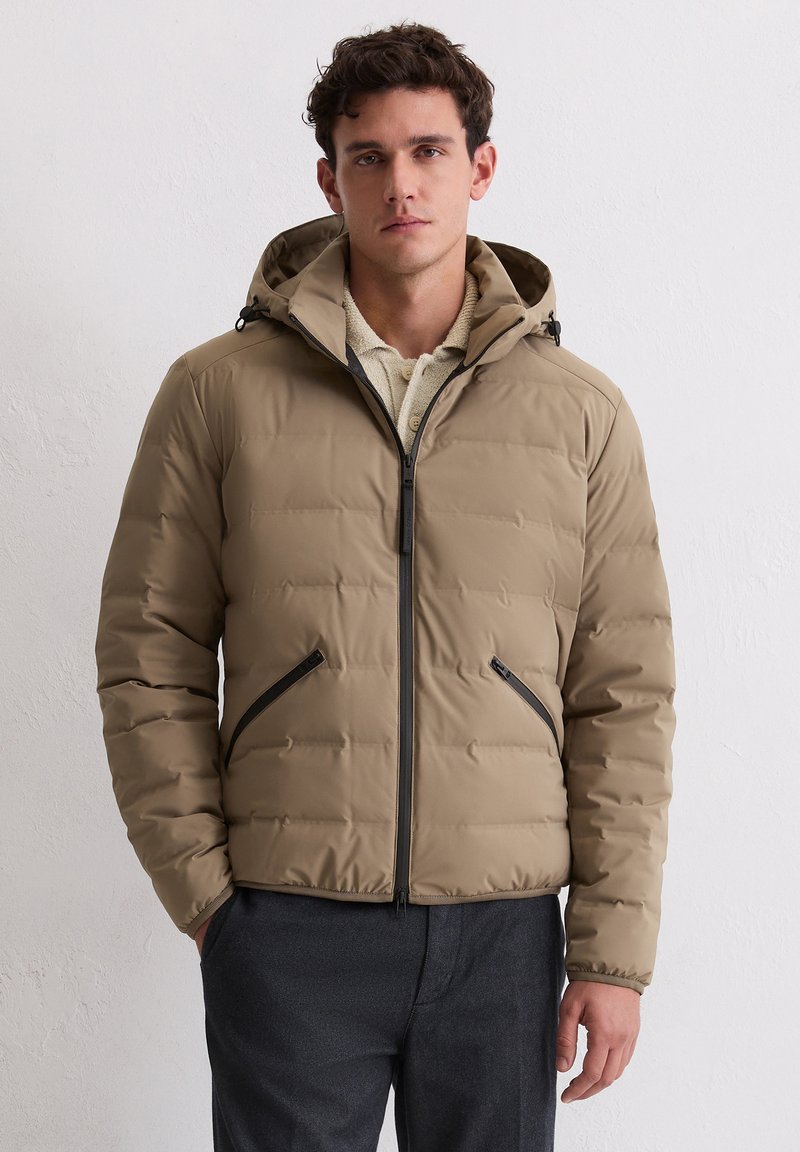 Hellbraune Steppjacke mit Kapuze, vorne mit Reißverschluss, zwei Reißverschlusstaschen und gesteppten Blöcken für Struktur. Schwarze Akzente an den Reißverschlüssen.