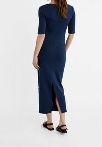 Granatowa dopasowana sukienka z krótkimi rękawami, długości do połowy łydki, z rozcięciem z tyłu i gładką teksturą. Noszona z czarnymi sandałami.