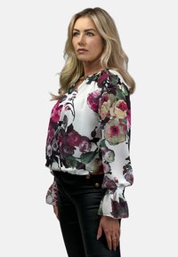 Kwiatowa bluzka z białym tłem i wzorami fioletowych, różowych oraz zielonych kwiatów. Tkanina jest miękka, z bufiastymi, rozkloszowanymi rękawami.