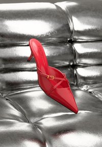 Rote spitze High-Heel-Pantoletten mit Kreuzband und einer kleinen Schnalle auf einer glänzenden silbernen gepolsterten Oberfläche.