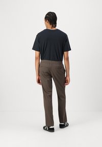 Svart t-skjorte med korte ermer, brune elastiske bukser med liv, og svarte sandaler med hvite detaljer, sett fra bakgrunnen mot en enkel bakgrunn.