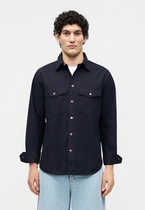 Άνδρας με σγουρά μαλλιά που φοράει σκούρο μπλε πουκάμισο με τσέπες στο στήθος και ανοιχτόχρωμα τζιν, στέκεται μπροστά σε ένα μονόχρωμο φόντο.